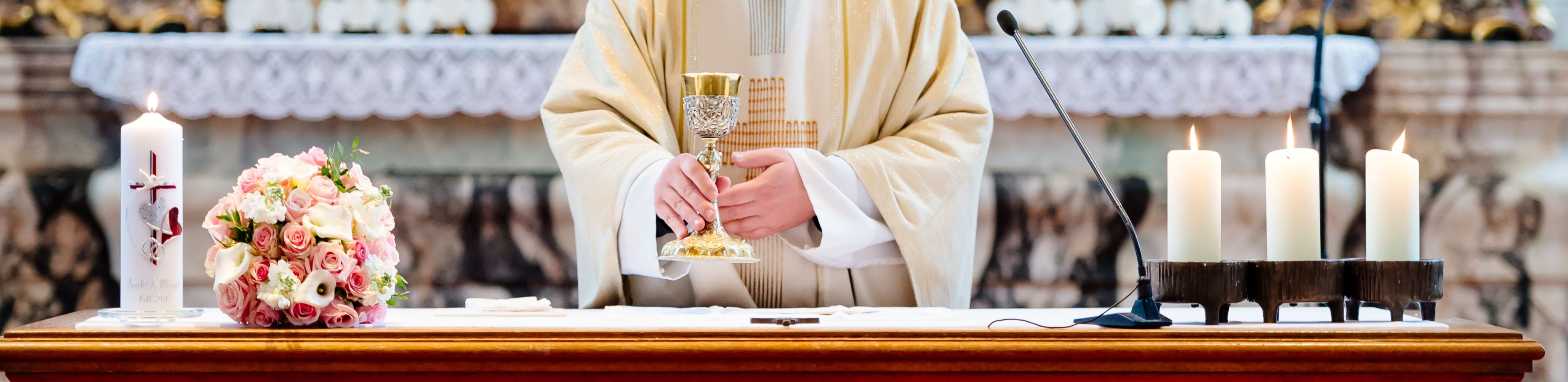 image of priest at altar celebrating mass, holding the ciborium, candles are lit on either side, and there is a bouquet of roses on the altar